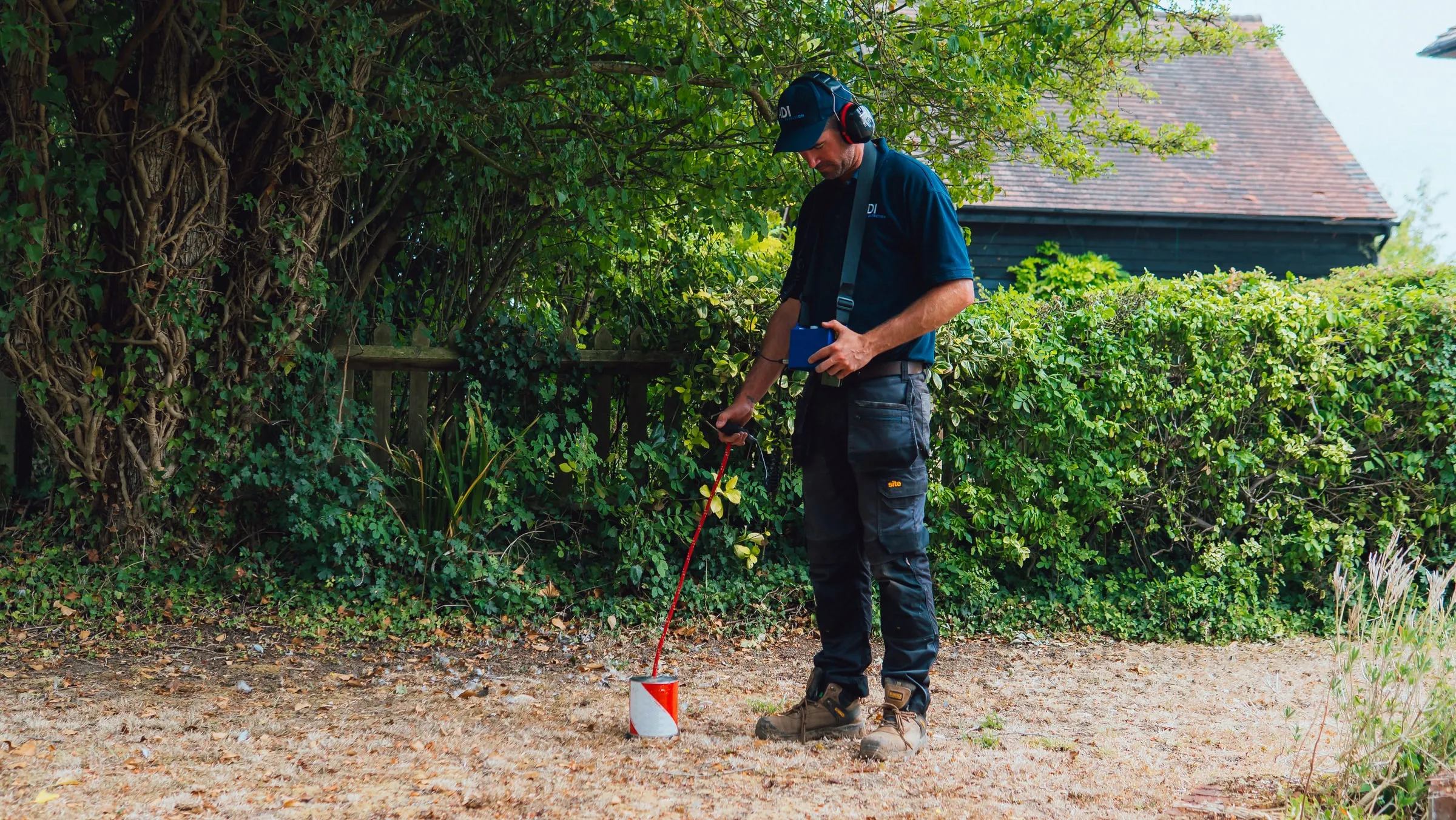 ADI engineer walking a garden with a tracer gas detector wand, a house roof visible behind the hedge
